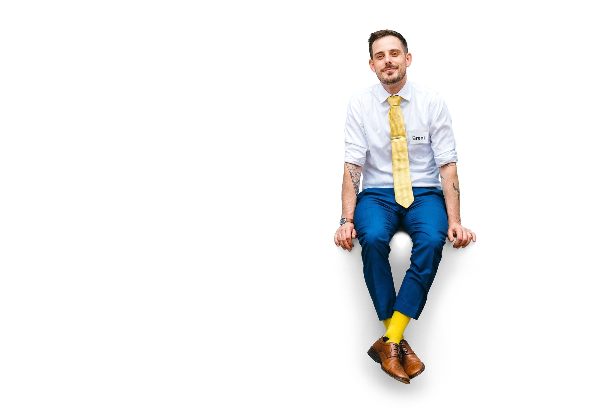 A picture of Brent, sitting on a the letters Helloooo! Wearing a white shirt, blue pants, and yellow tie and socks, with a name tag that says "Brent".
