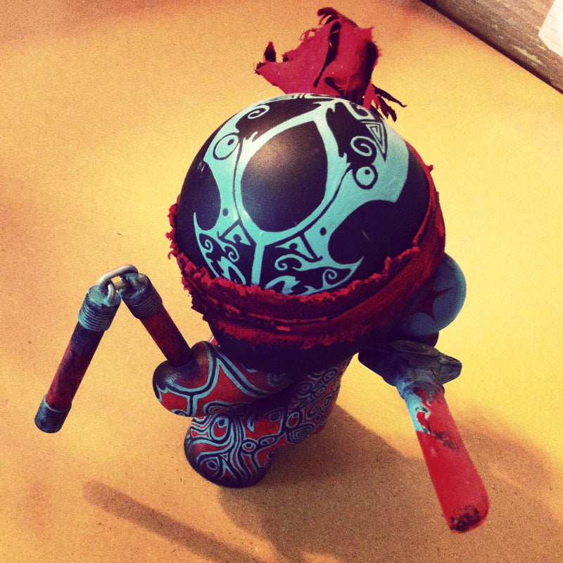 Custom-painted Kidrobot Munny DIY vinyl art figure. It features a tribal and traditional tattoo-inspired aesthetic in red and light blue colors.The figure is depicted wielding a toy nunchaku and sword with a blood-like stain.